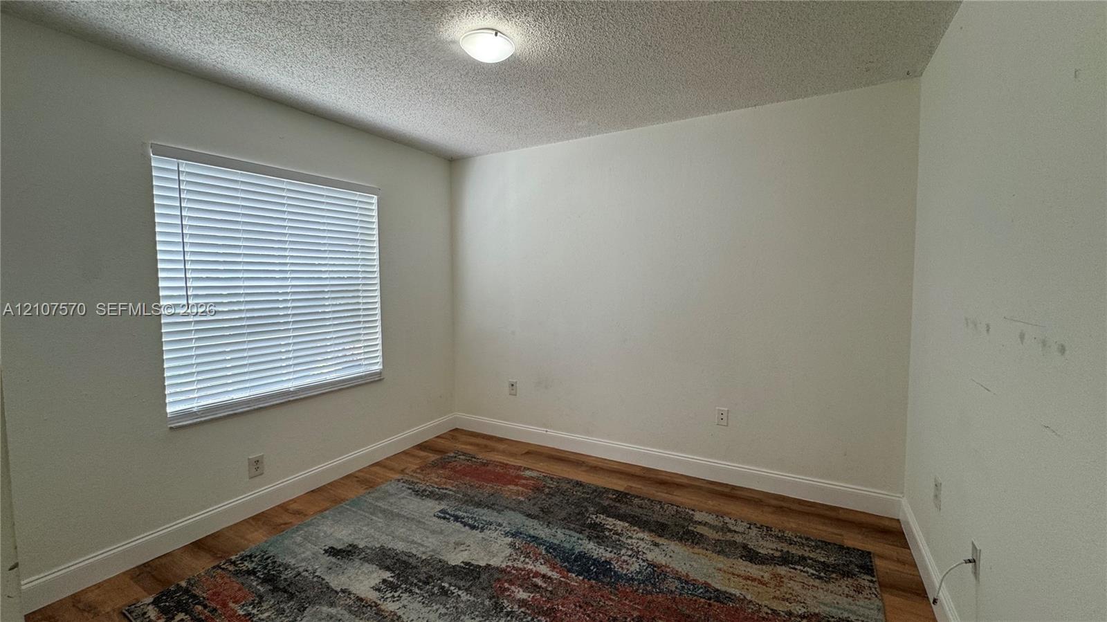 Neutral-toned interior room featuring wood-finish flooring, a single window with horizontal blinds, and a central ceiling-mounted light fixture