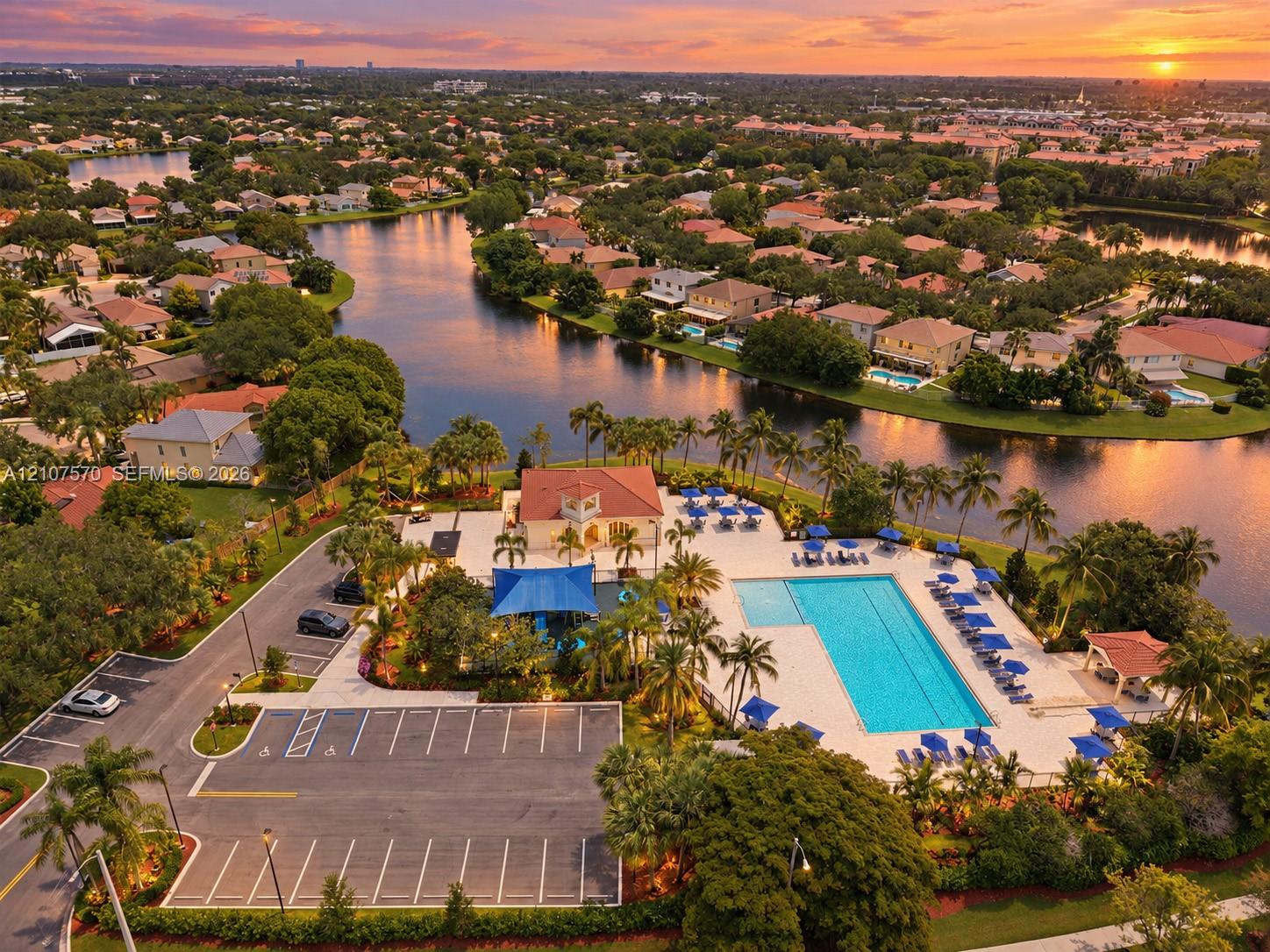 Aerial perspective of the community recreation facility featuring a swimming pool, paved parking area, clubhouse building, and mature palm trees situated along the waterfront