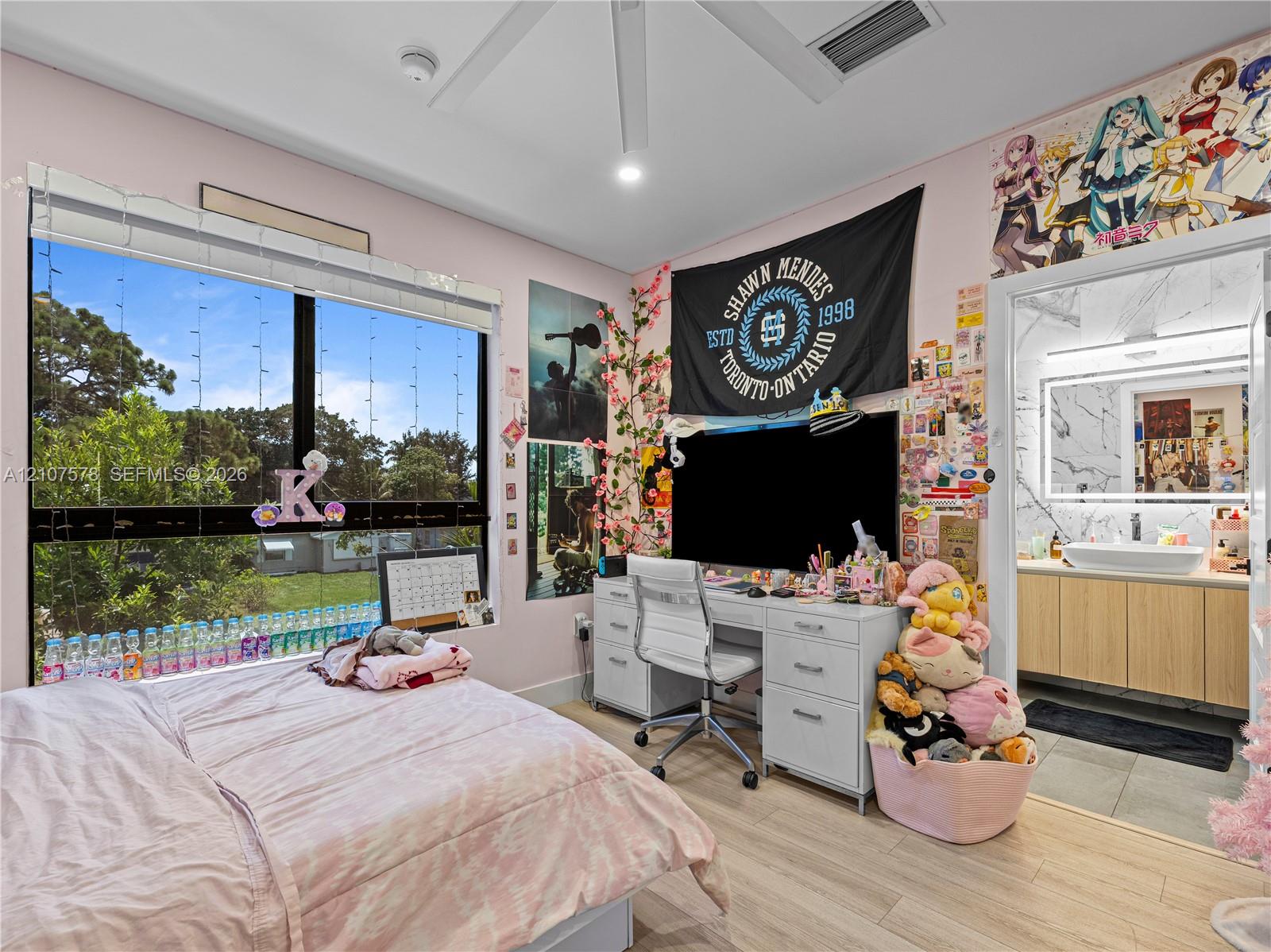 Large multi-pane window, wood-finish flooring, ceiling fan with integrated lighting, and en-suite bathroom featuring a light-wood vanity with a white vessel sink and backlit mirror
