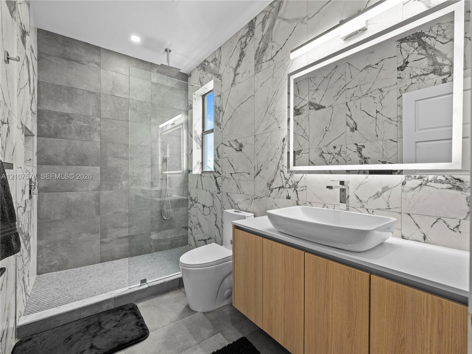 Bathroom featuring large-format wall-to-wall stone-style tile, a walk-in glass shower enclosure with a rainfall showerhead, a wood-finish vanity with a rectangular vessel sink, and a large illuminated vanity mirror