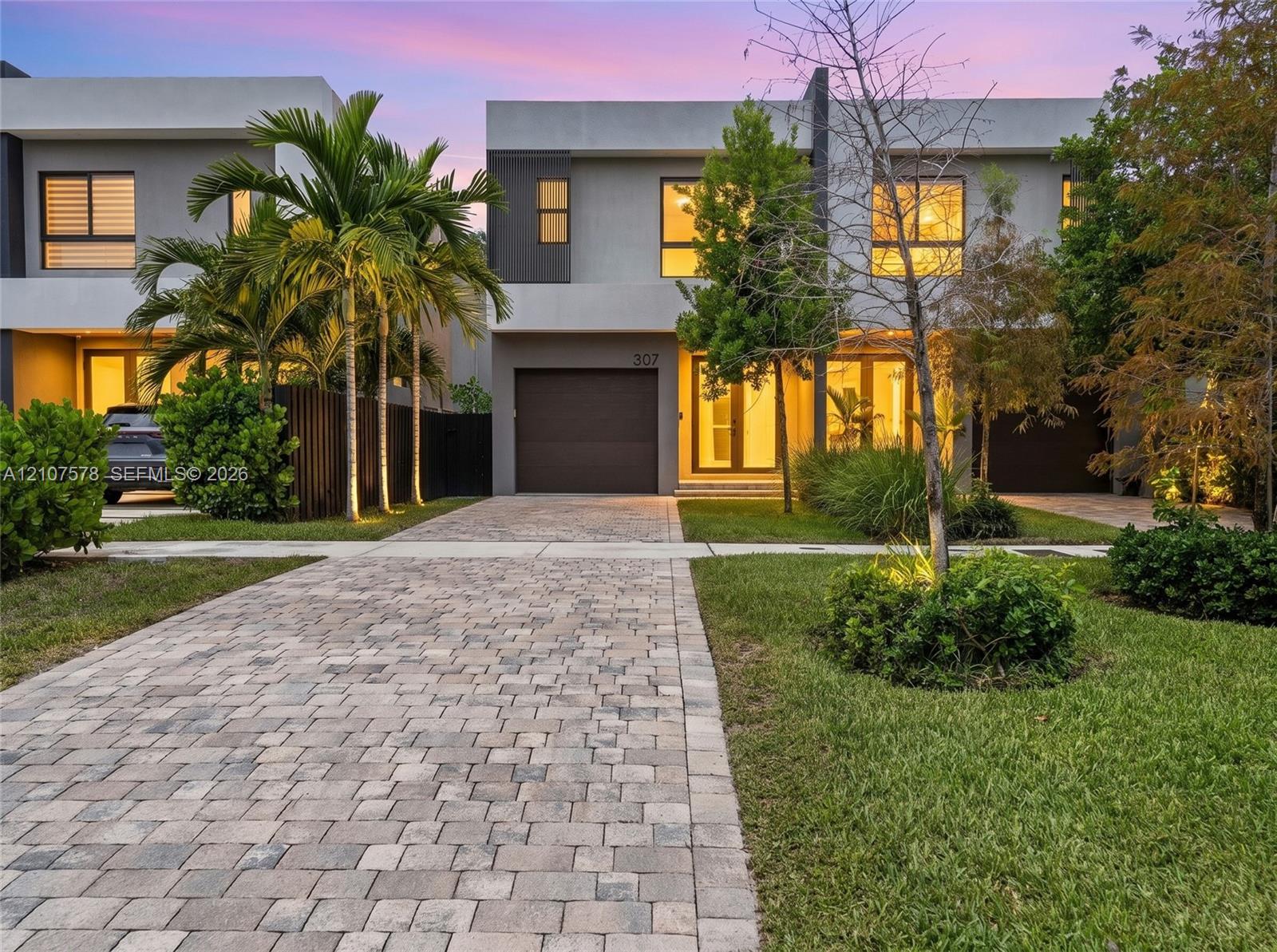 Modern architectural exterior featuring a concrete facade, paver driveway, integrated garage, and floor-to-ceiling glass entry doors