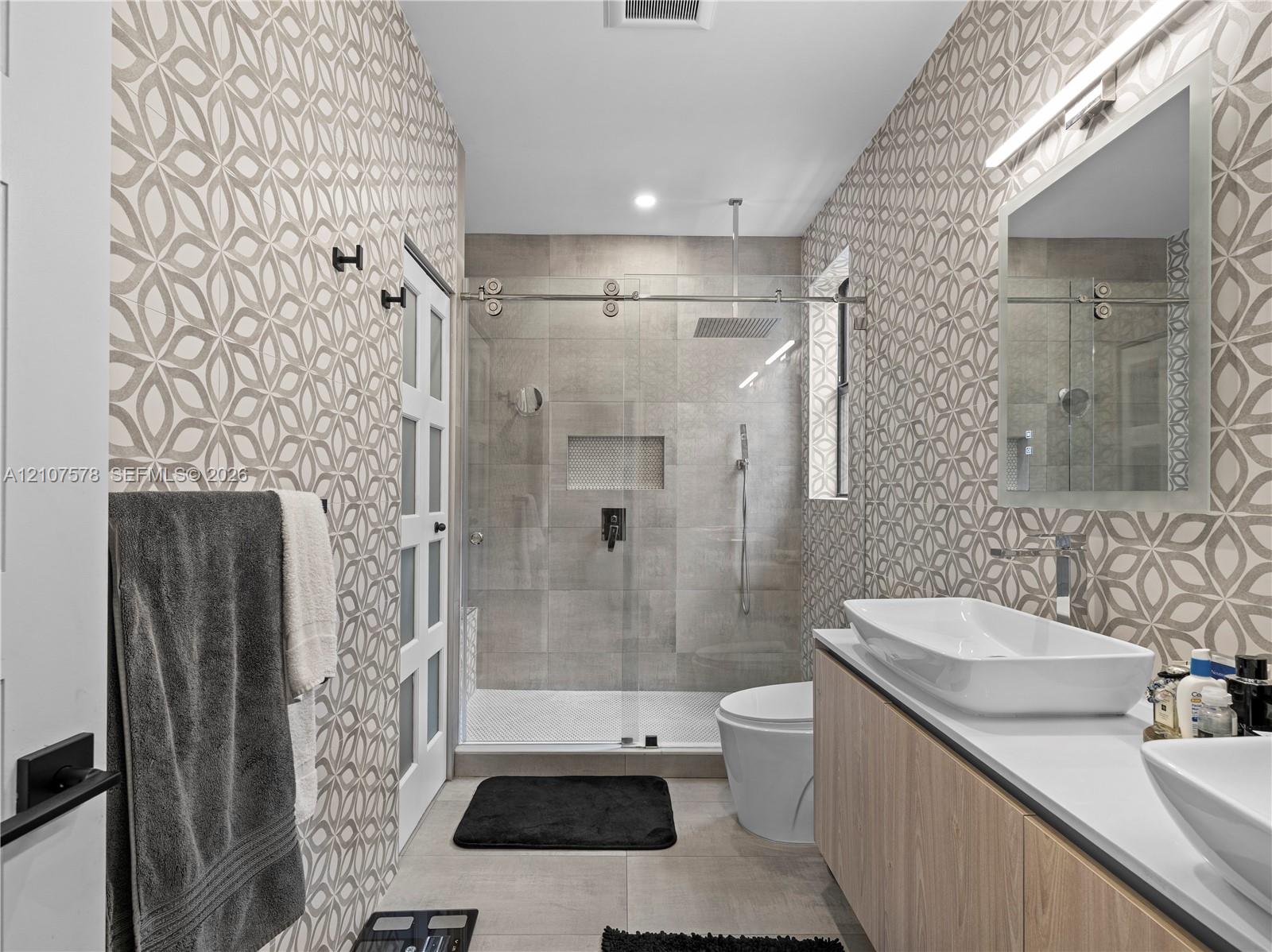 Floating wood-finish vanity with dual vessel sinks, walk-in shower with glass enclosure and rain-style showerhead, patterned wall tile, and built-in recessed shower niche