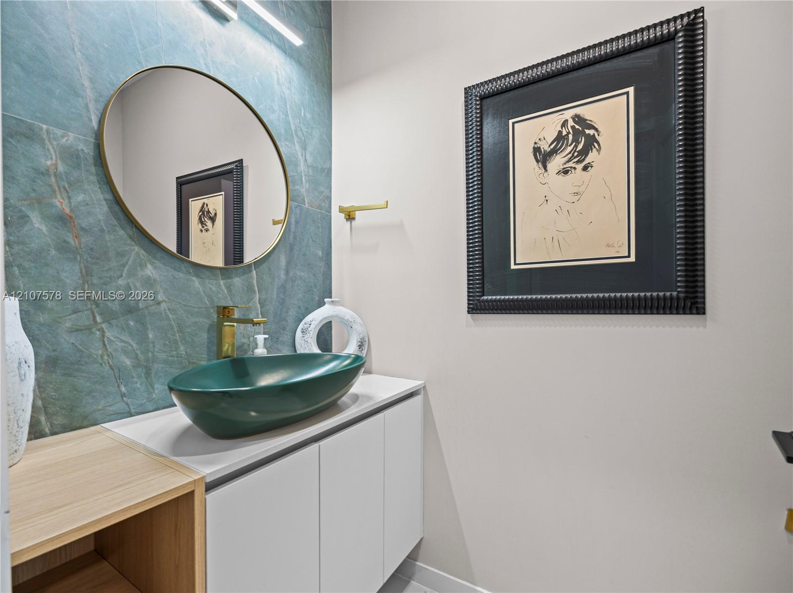 Floating vanity with white cabinetry and wood-finish shelving, topped with a green vessel sink, gold-tone faucet, and large-format teal tile backsplash featuring an oval mirror and minimalist linear lighting