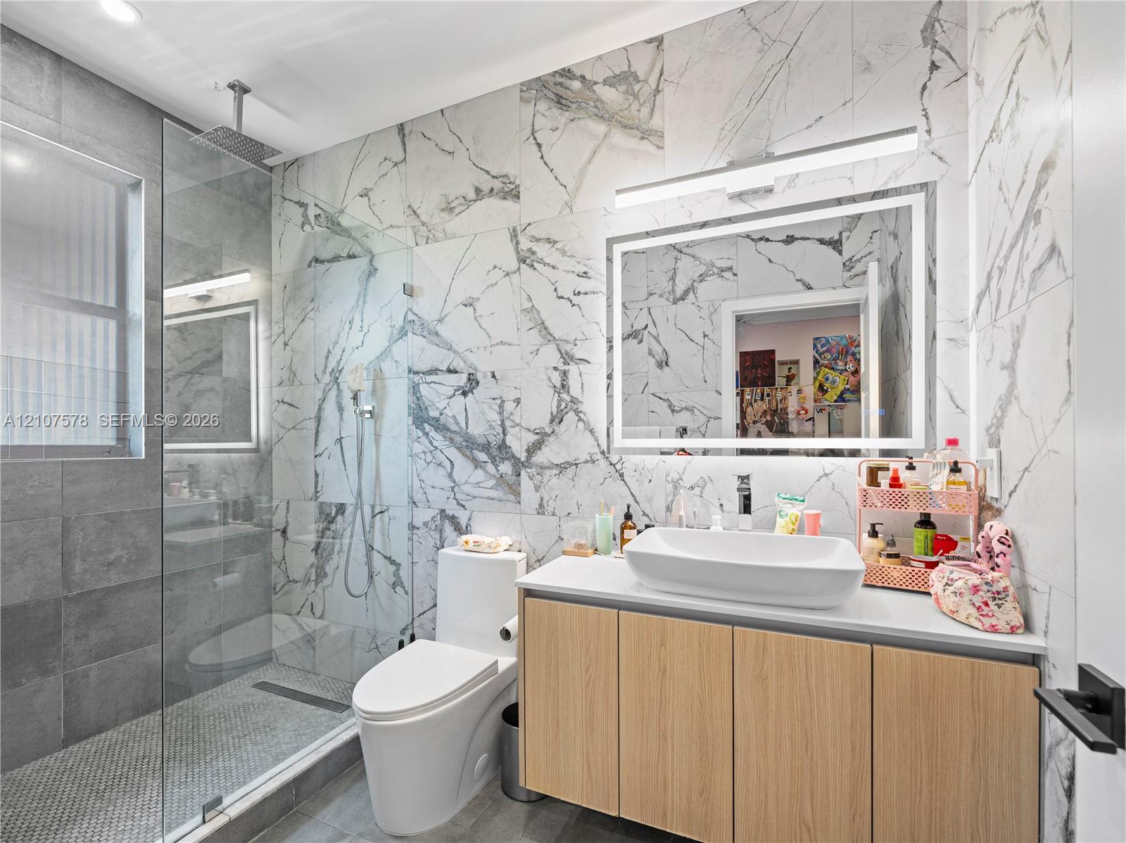 Bathroom featuring floor-to-ceiling large-format veined tile, walk-in shower with a glass enclosure and rainfall showerhead, floating vanity with a vessel sink, and an integrated LED-backlit mirror