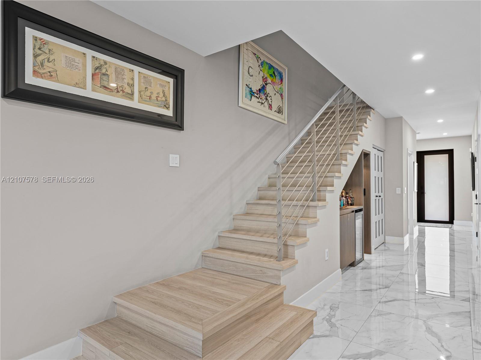 Staircase featuring wood-finish treads, metal horizontal railing, light-toned stone-finish flooring, recessed ceiling lighting, and built-in under-stair wet bar cabinetry