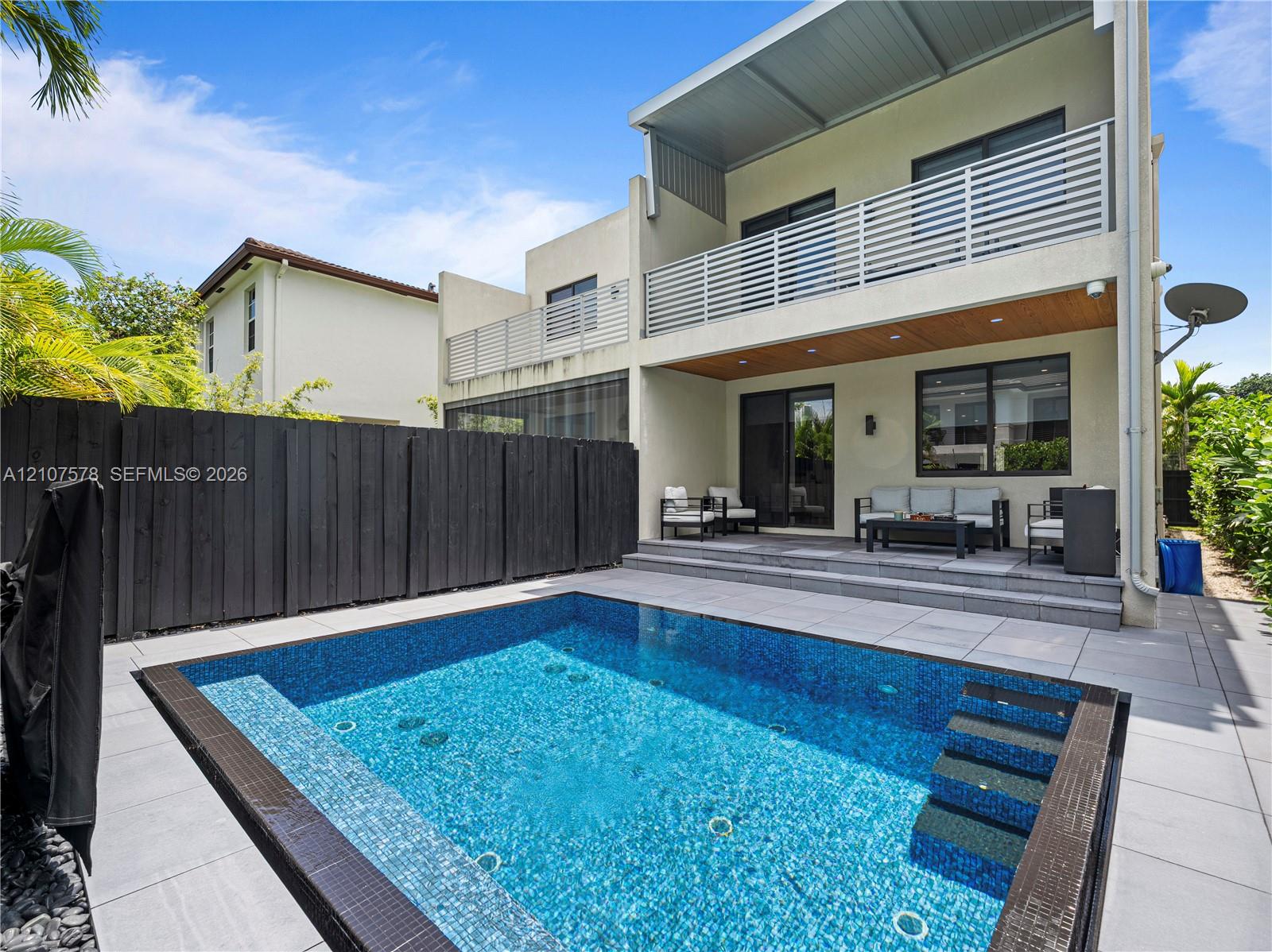 Exterior space featuring a rectangular in-ground pool with mosaic tile interior, light-toned stone paver decking, a covered rear patio with wood-finish ceiling, and a horizontal slat privacy fence
