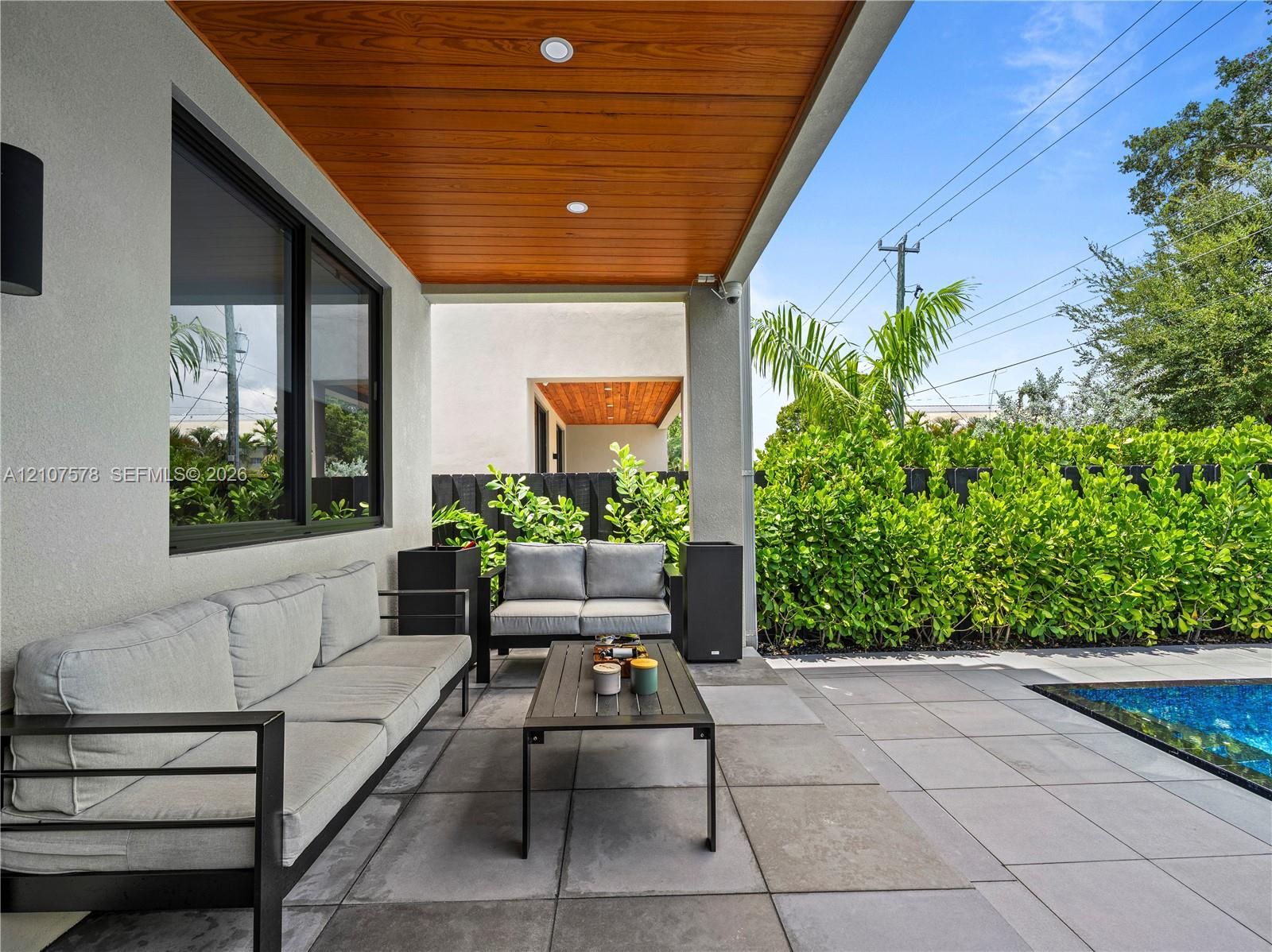 Covered patio featuring a wood-plank ceiling, recessed lighting, and large-format porcelain paver flooring, complemented by a swimming pool and perimeter privacy hedge