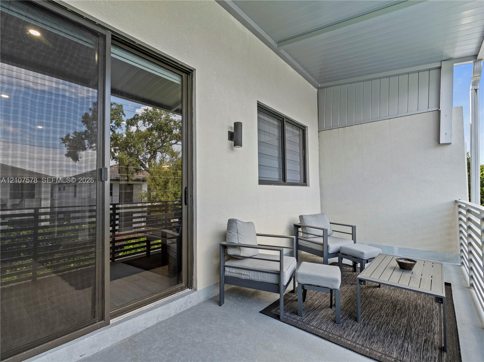 Covered balcony featuring sliding glass doors, horizontal slat railings, neutral stucco exterior wall, and wall-mounted exterior lighting