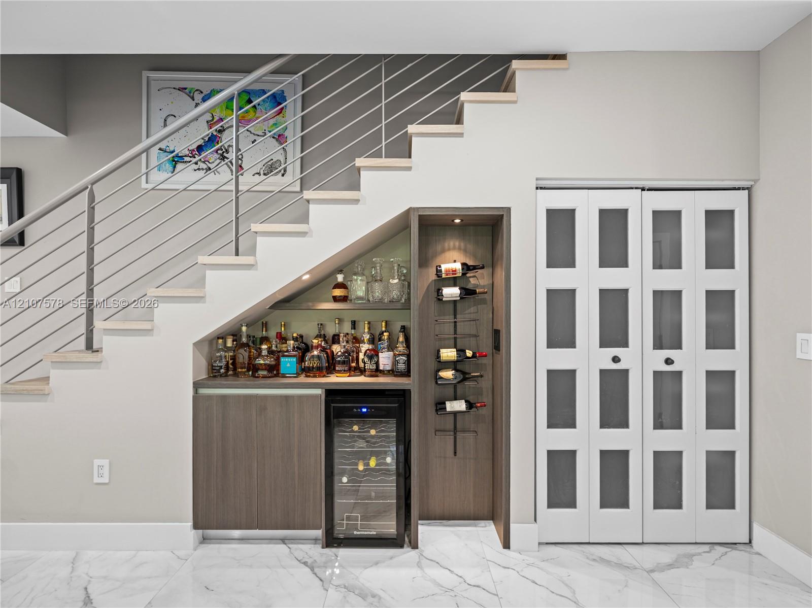 Built-in wet bar featuring wood cabinetry, integrated wine refrigerator, vertical wall-mounted wine rack, and recessed accent lighting, positioned beneath a staircase with metal railing and finished with stone-style tile flooring