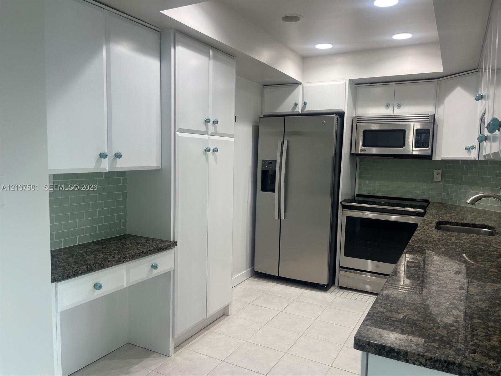 White cabinetry with decorative knobs, stainless steel appliances, dark stone-finish countertops, tiled backsplash, and recessed lighting