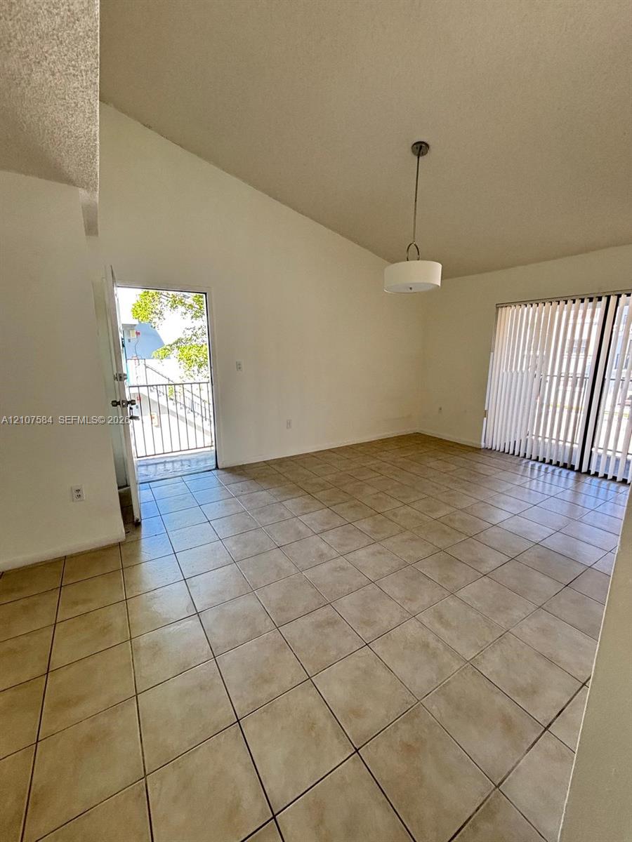 Vaulted ceiling, neutral tile flooring, drum-style pendant lighting, and sliding glass doors with vertical blinds