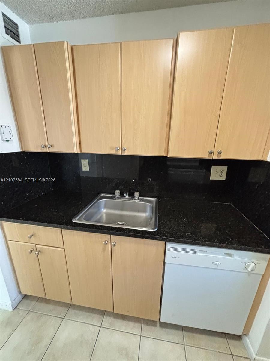 Kitchen area featuring light wood-finish cabinetry, dark solid-surface countertops, a stainless steel sink with a dual-handle faucet, an integrated dishwasher, and ceramic tile flooring