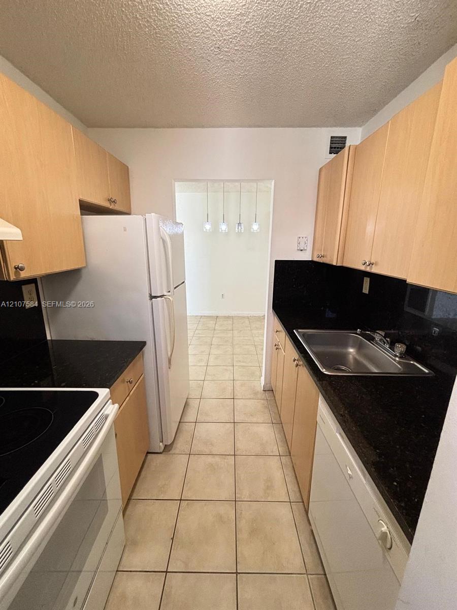 Galley kitchen featuring light-toned wood-finish cabinetry, dark solid-surface countertops, ceramic tile flooring, stainless steel sink, and integrated appliances