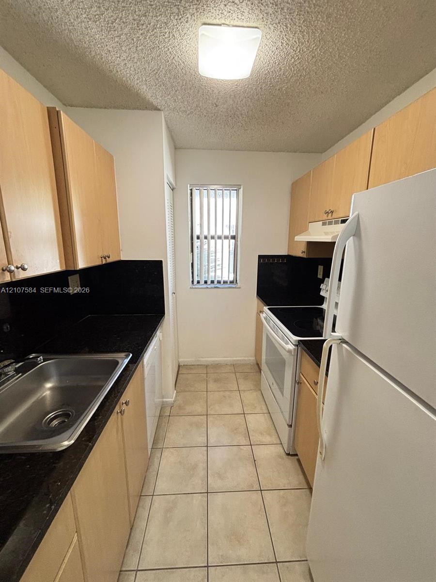 Galley kitchen featuring light-toned cabinetry, dark solid-surface countertops, ceramic tile flooring, and stainless steel sink