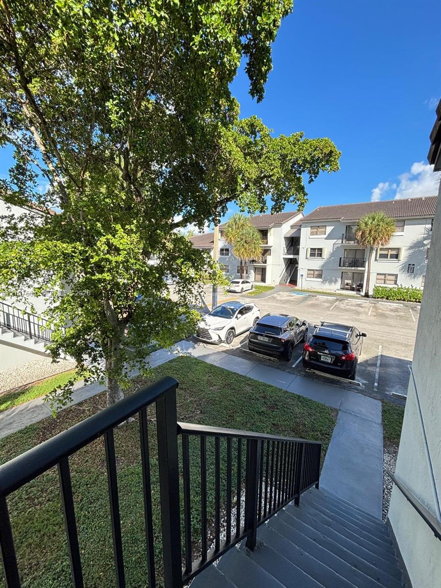 Exterior staircase featuring black metal railings, paved parking area, mature landscaping, and multi-story building exterior