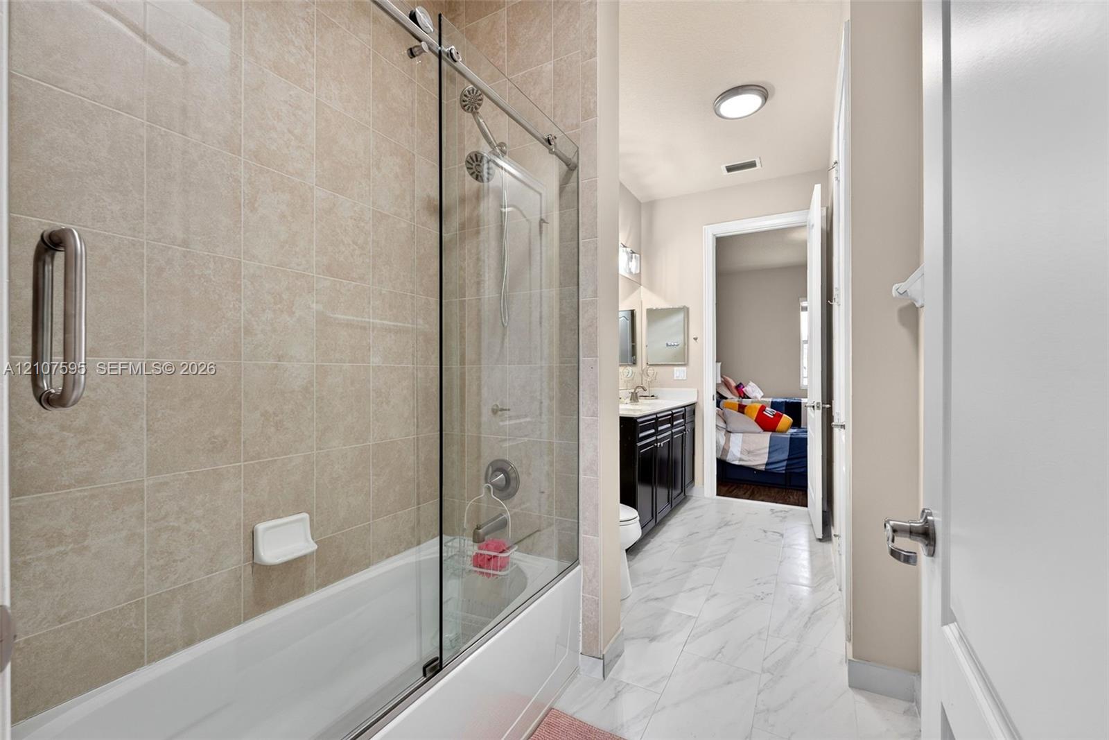 Bathroom featuring a tub-shower combination with floor-to-ceiling neutral tile surround and glass sliding doors, dark wood-finish cabinetry, stone-style vanity countertop, and white marble-finish tile flooring