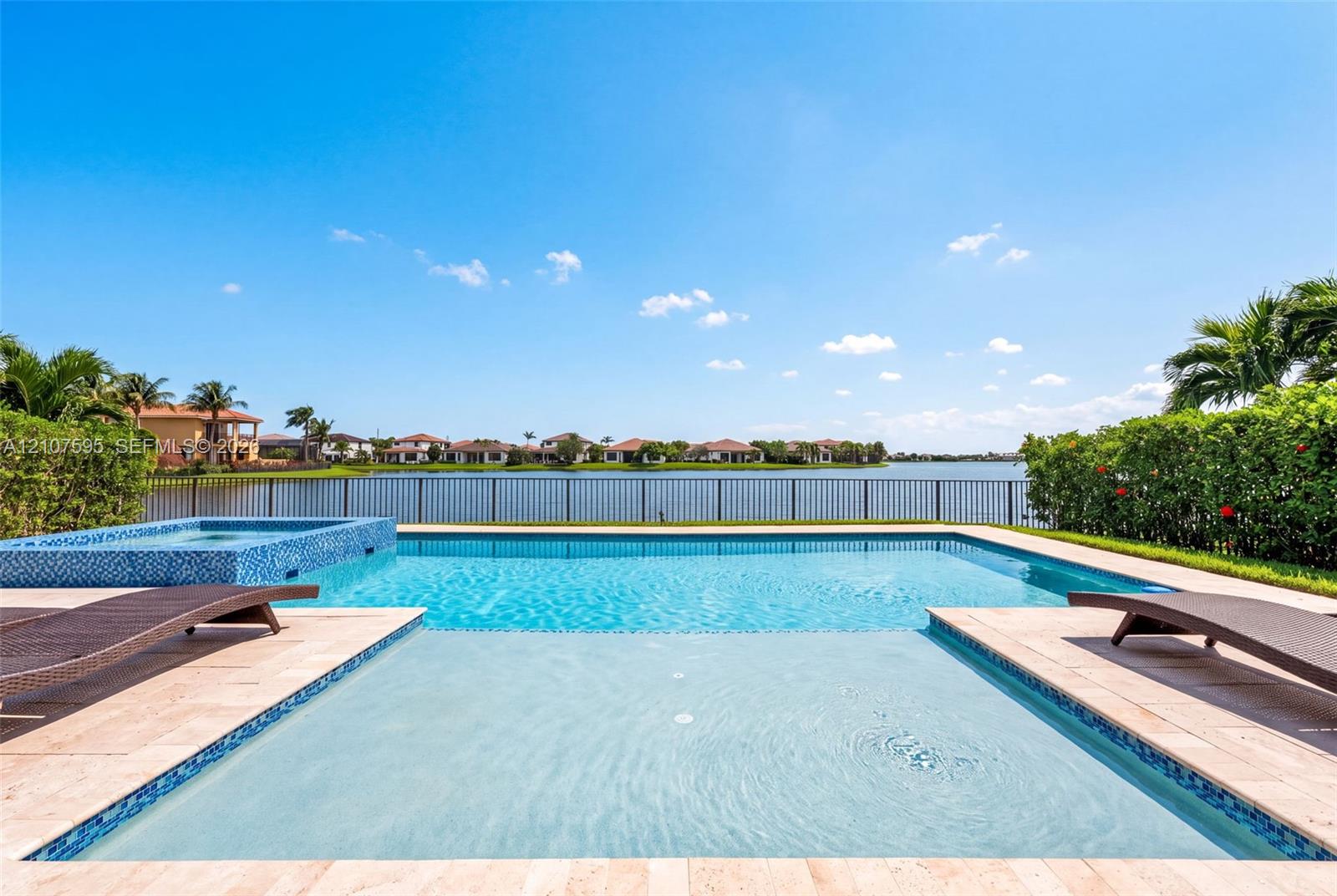 In-ground swimming pool and elevated spa with blue tile accents, stone-finish decking, and metal perimeter fencing overlooking a lake