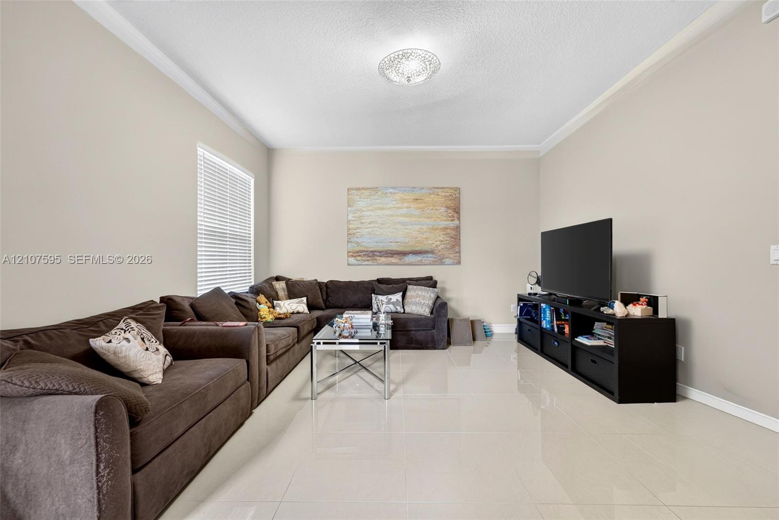 Spacious room featuring neutral wall tones, large-format tile flooring, crown molding, and a central ceiling-mounted light fixture