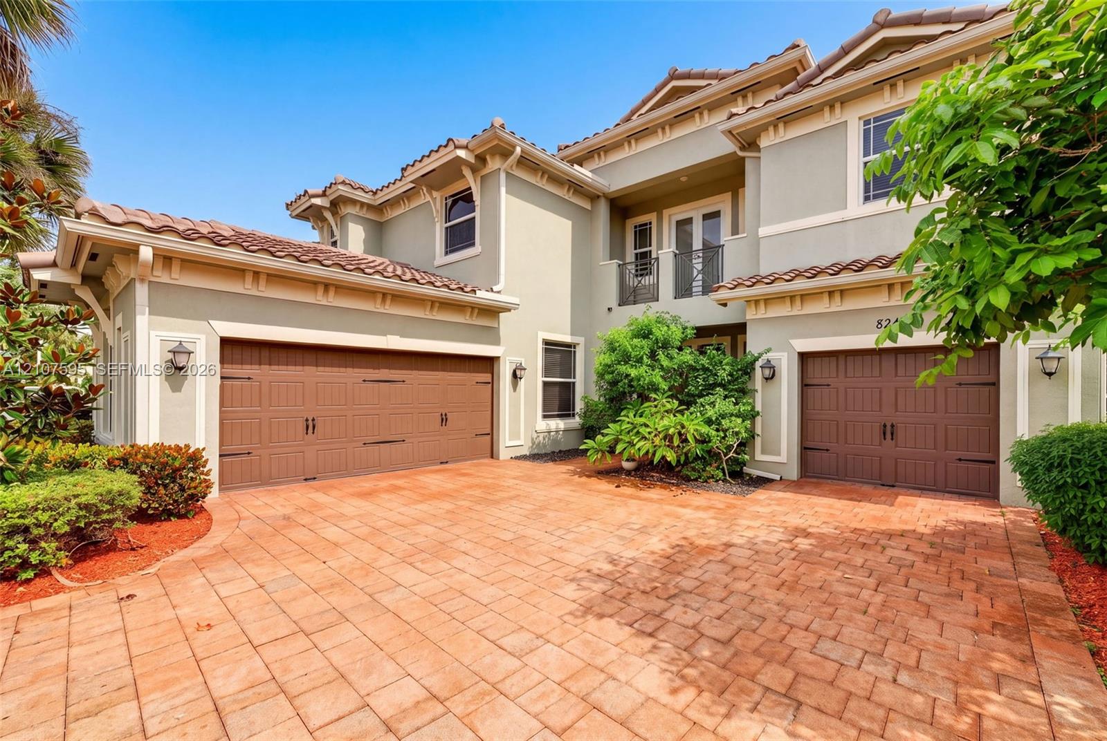 Stucco exterior featuring a tile roof, brick-style paver driveway, two-car garage, and upper-level balcony with metal railing