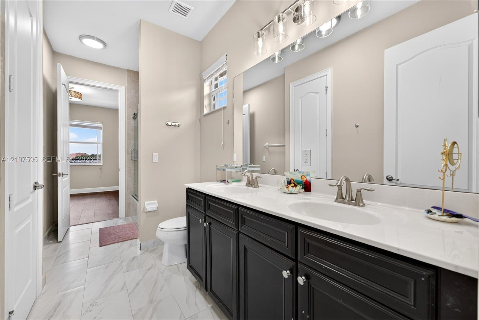 Double vanity with dark cabinetry, stone-finish countertops, dual under-mount sinks, and large wall-mounted mirror, featuring porcelain tile flooring and multi-bulb task lighting