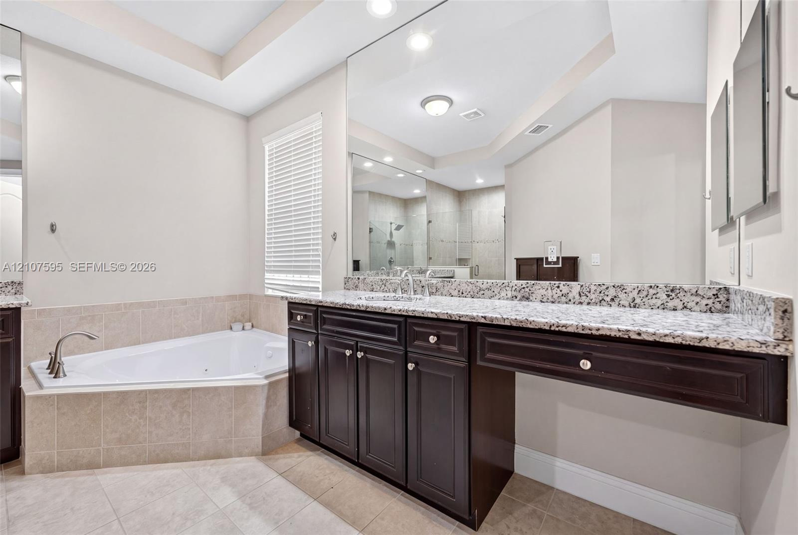Built-in dark wood vanity featuring a speckled stone countertop, large-scale wall-mounted mirror, corner jetted soaking tub with tile surround, and neutral tile flooring
