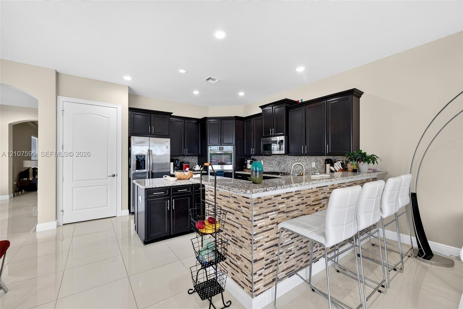 Kitchen featuring dark cabinetry, stone countertops, a tile-accented breakfast bar, stainless steel appliances, and large-format tile flooring