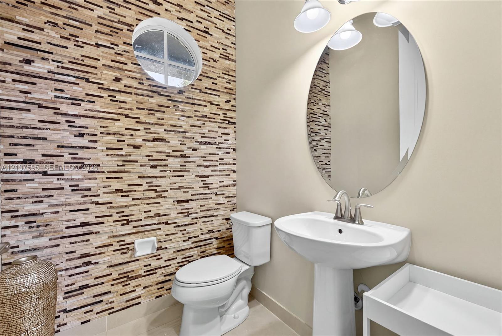 Stacked stone accent wall, oval wall-mounted mirror, white pedestal sink with polished nickel faucet, and circular window