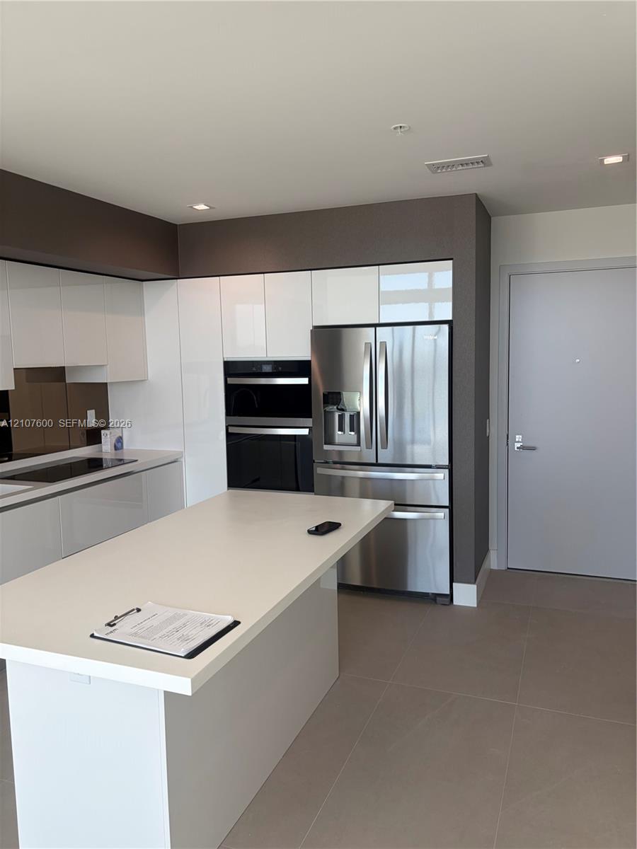 Kitchen featuring a central island with a solid surface countertop, high-gloss white cabinetry, stainless steel appliances including a built-in wall oven, and ceramic tile flooring