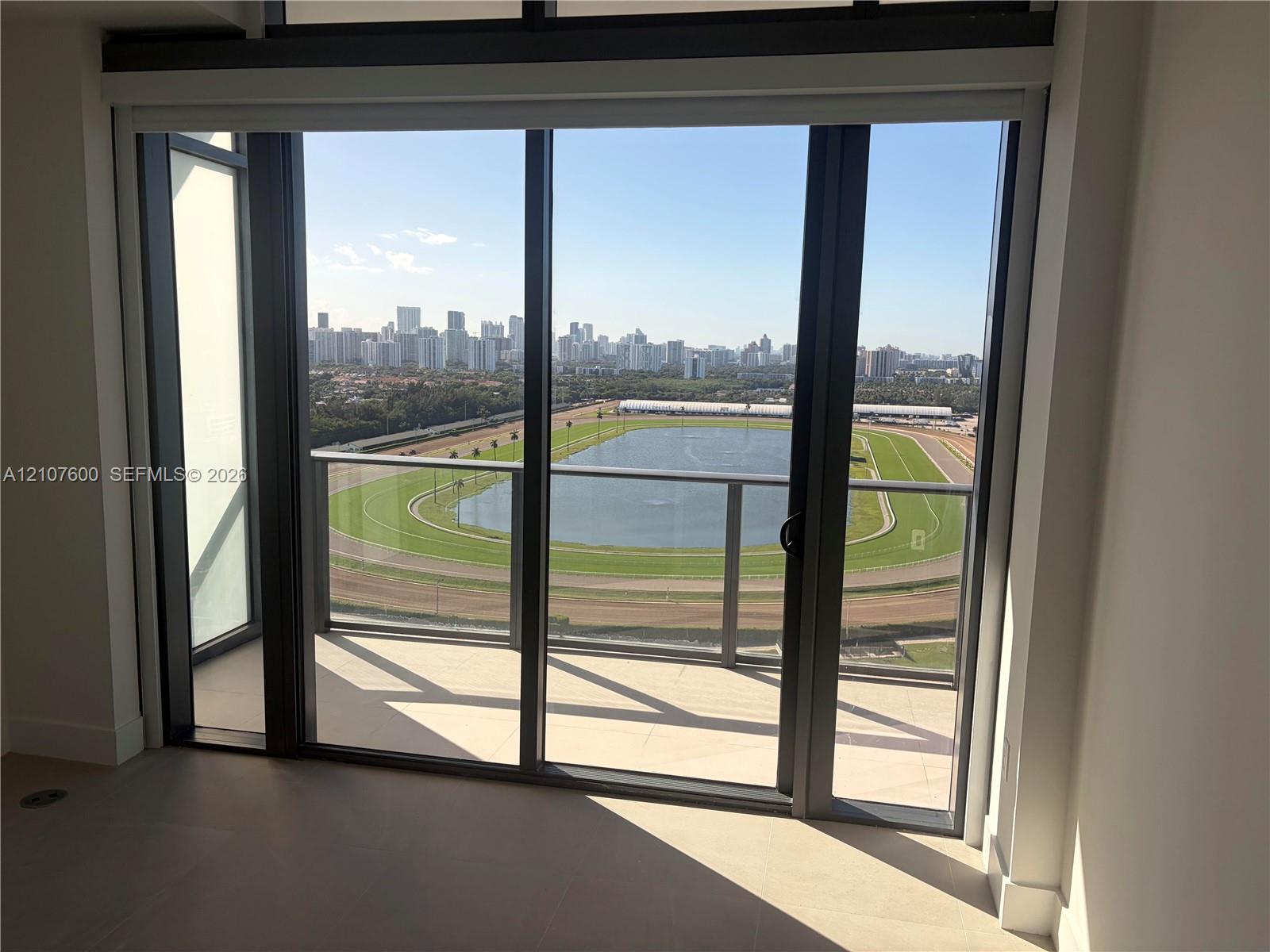 Floor-to-ceiling glass sliding doors opening to a covered balcony with a glass panel railing, tile flooring, and expansive panoramic scenery of a racetrack, lake, and urban skyline