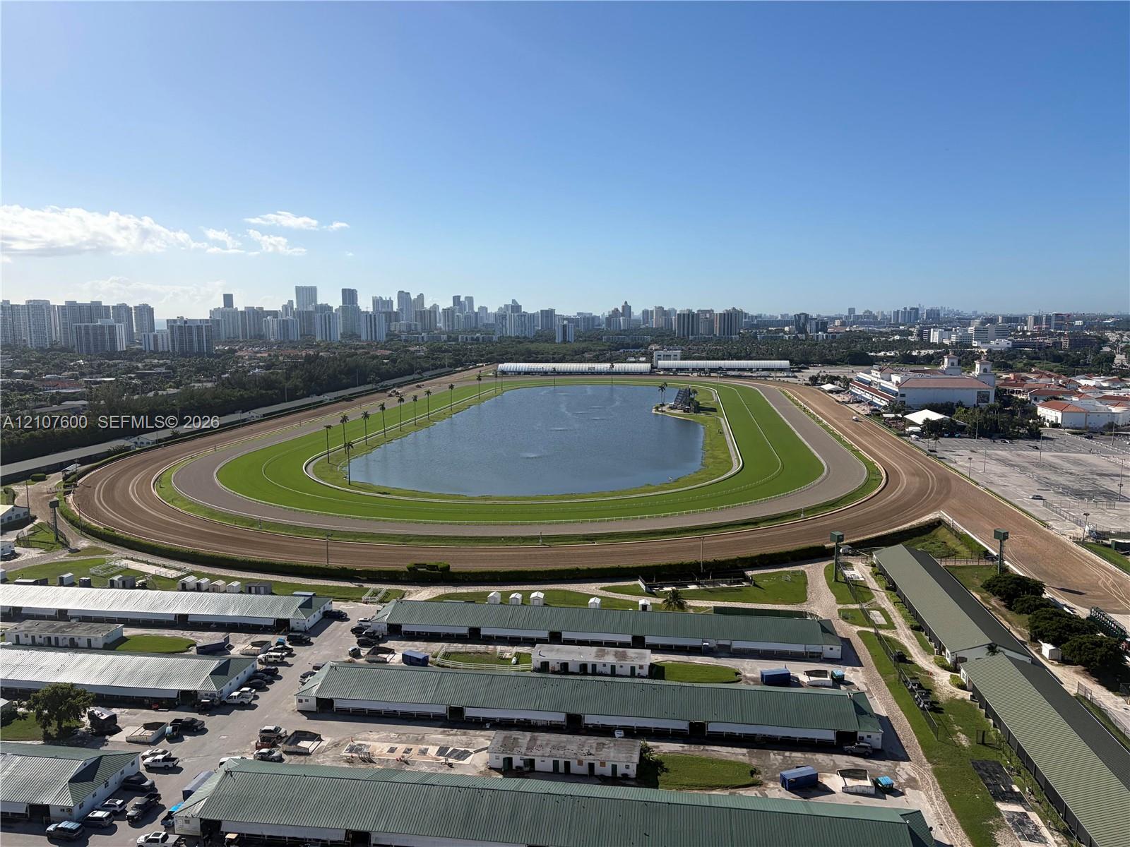 Aerial perspective of a large-scale track facility featuring a central grassed oval, an expansive interior water feature, a multi-lane dirt track, and rows of metal-roofed structures Aerial perspective of a large-scale track facility featuring a central grassed oval, an expansive interior water feature, a multi-lane dirt track, and rows of metal-roofed structures