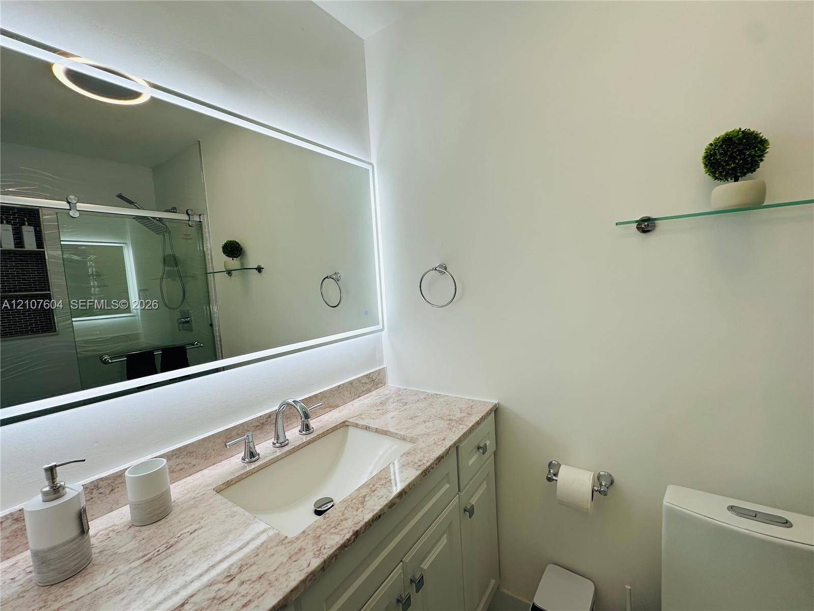 Bathroom vanity featuring a stone countertop with an undermount sink, widespread metal faucet, wall-mounted backlit mirror, and glass shelf