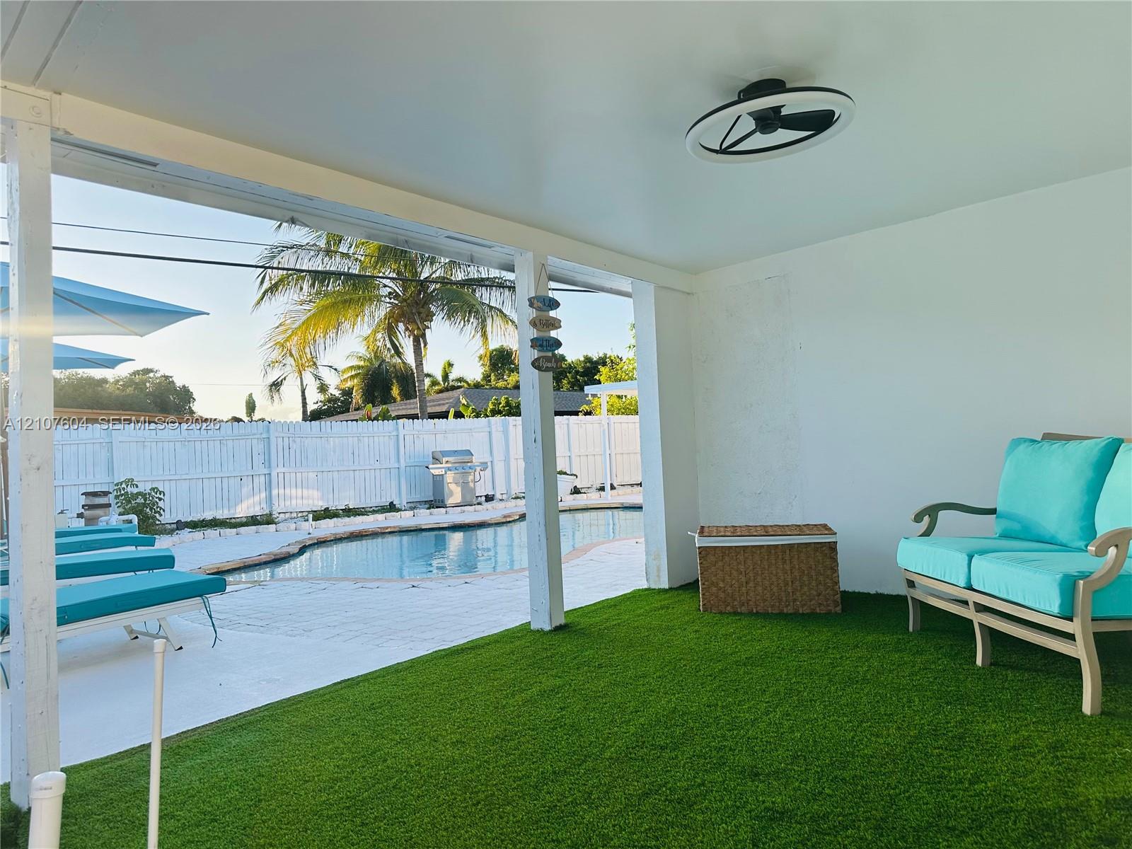 Covered patio featuring artificial turf flooring, ceiling-mounted light fixture, white structural pillars, pool area, and white perimeter fencing