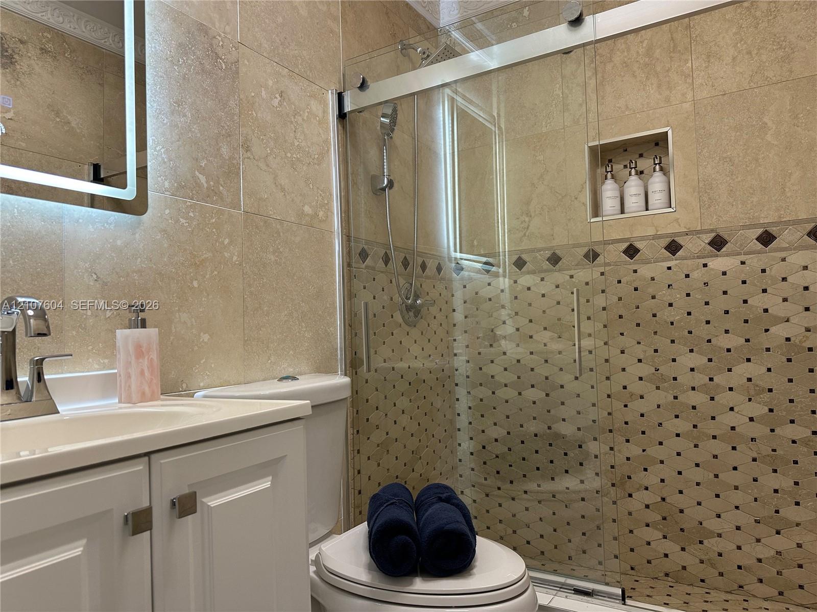 Bathroom featuring a vanity with a white cabinet, stone-tile wall surfacing, a walk-in shower with a glass sliding door, a recessed shower niche, and a wall-mounted illuminated mirror