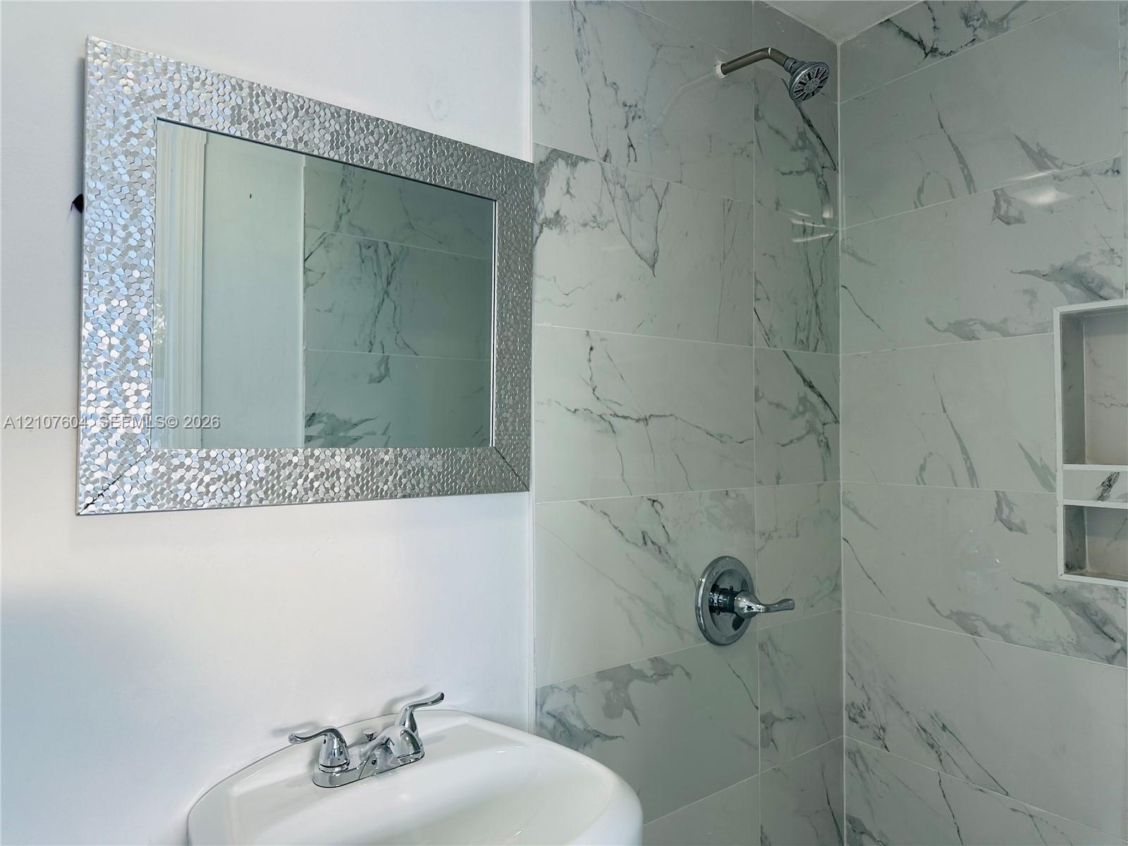 Wall-mounted bathroom vanity featuring a basin sink with a two-handle chrome faucet, accompanied by a rectangular decorative-frame mirror and a walk-in shower with large-format stone-patterned tile, chrome hardware, and an integrated recessed soap niche