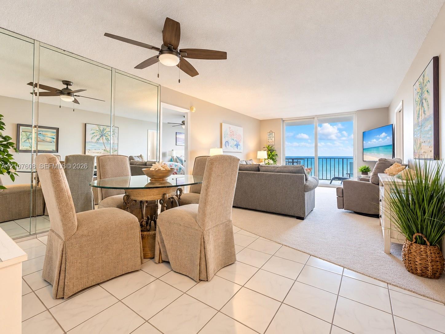 Open-concept living space featuring ceramic tile flooring, full-wall mirrored closet, ceiling fan with integrated lighting, and floor-to-ceiling sliding glass doors opening to an ocean backdrop