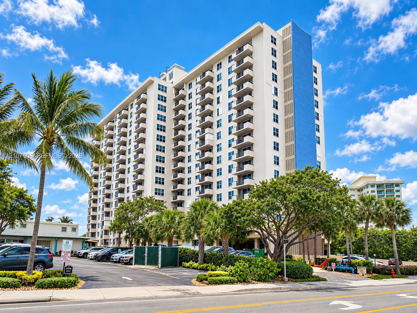 High-rise residential exterior featuring stacked private balconies, a distinctive vertical blue facade accent, and paved surface parking framed by mature palm trees and established landscaping