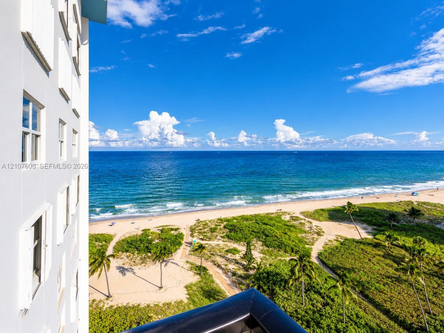 High-rise building exterior featuring a balcony with a black railing, overlooking an expansive coastal shoreline, sandy beach, and native vegetation