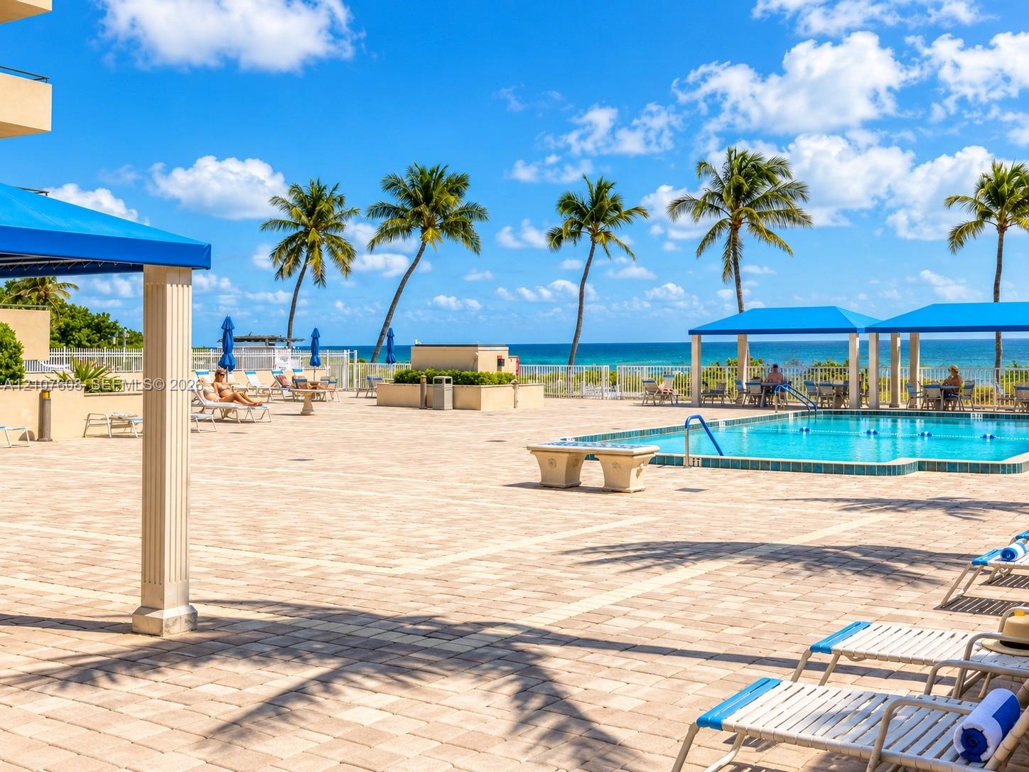 Paver pool deck with a swimming pool, shaded structural pavilions, and ocean backdrop