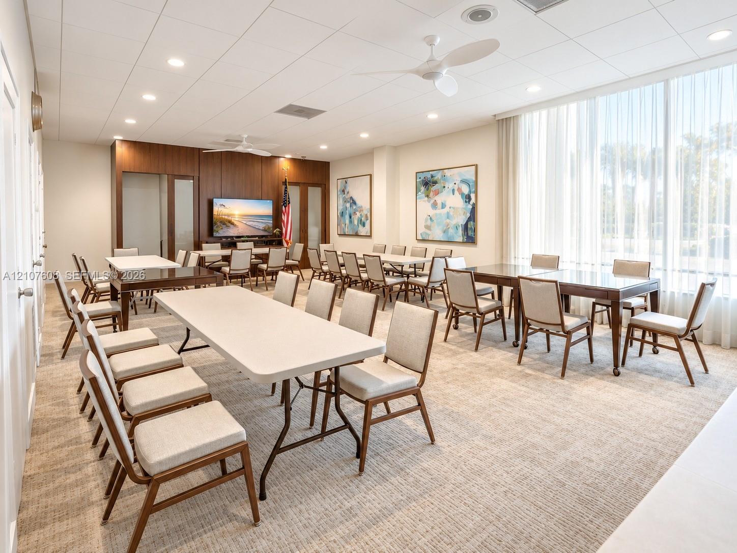 Spacious common room featuring floor-to-ceiling windows with sheer drapery, recessed ceiling lighting, dual ceiling fans, wood-paneled accent wall, and neutral carpeting