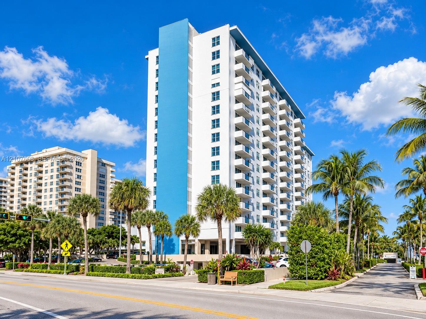 High-rise residential building exterior featuring a white and blue facade, prominent stacked balconies, and mature palm tree landscaping