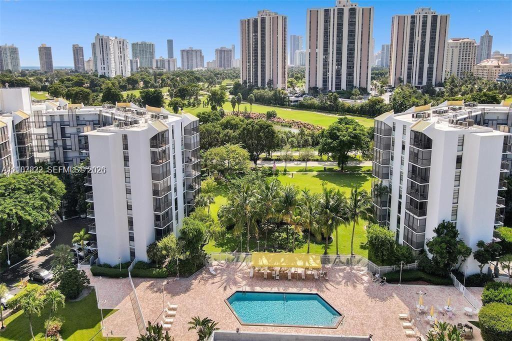 Aerial perspective of a multi-story residential complex featuring a paved pool deck, geometric swimming pool, landscaped lawn, and mature palm trees