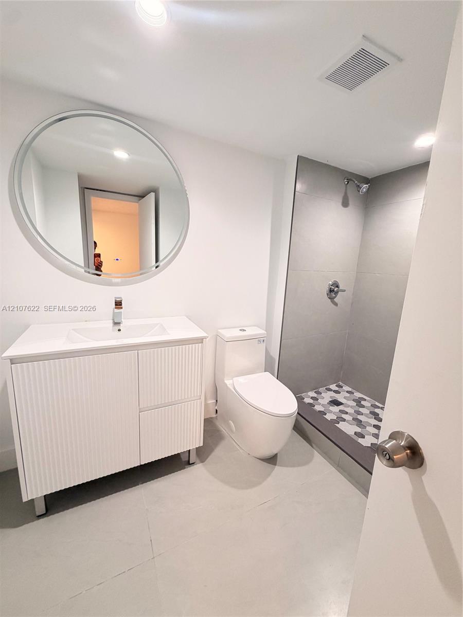 Bathroom featuring a white fluted vanity with a rectangular sink, a large circular mirror, a dual-flush toilet, and a walk-in shower with large-format grey wall tile and geometric mosaic floor tile