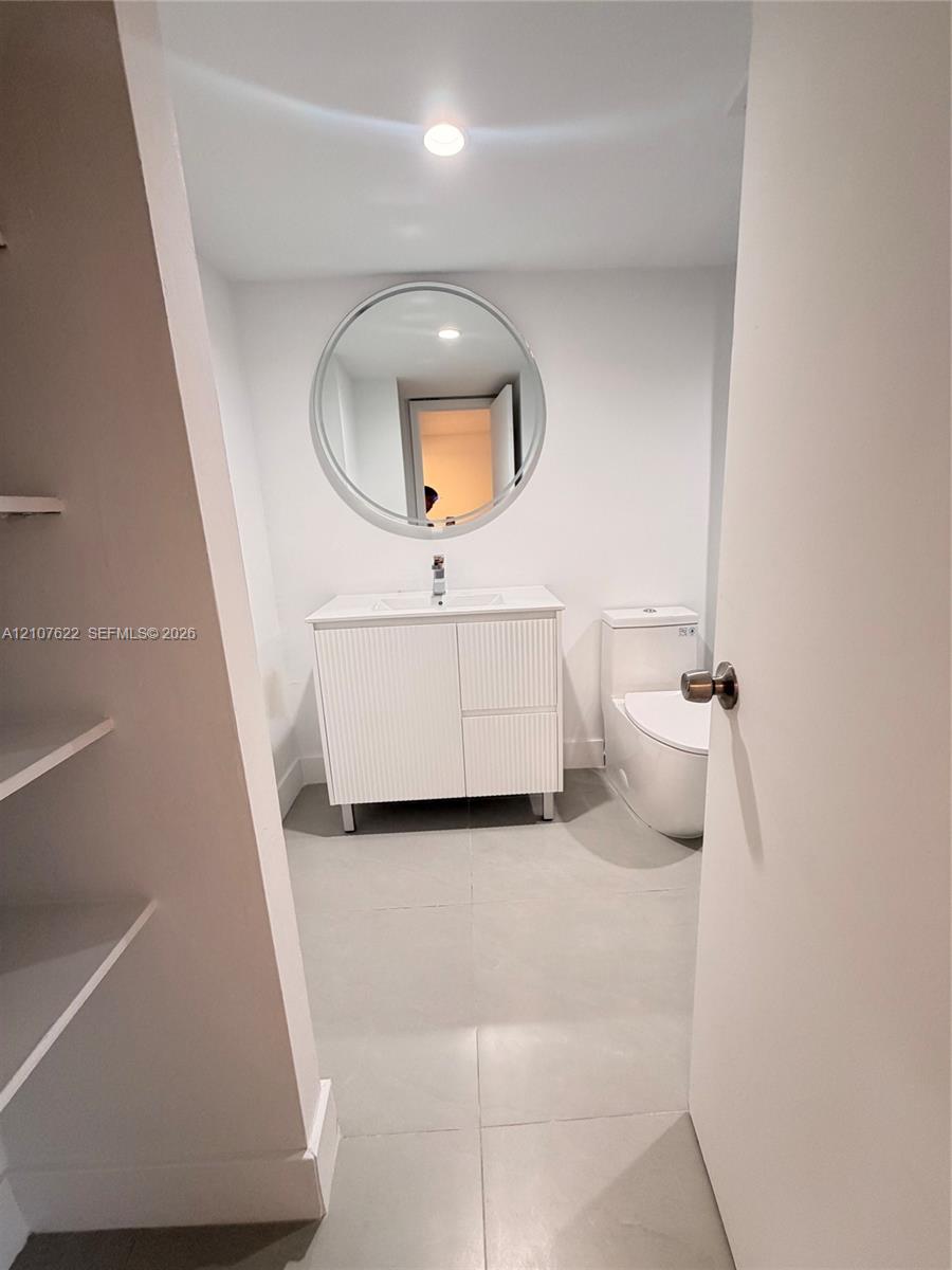 Bathroom featuring a ribbed-texture vanity, round mirror, white countertop with integrated sink, large-format tile flooring, and a skirted toilet