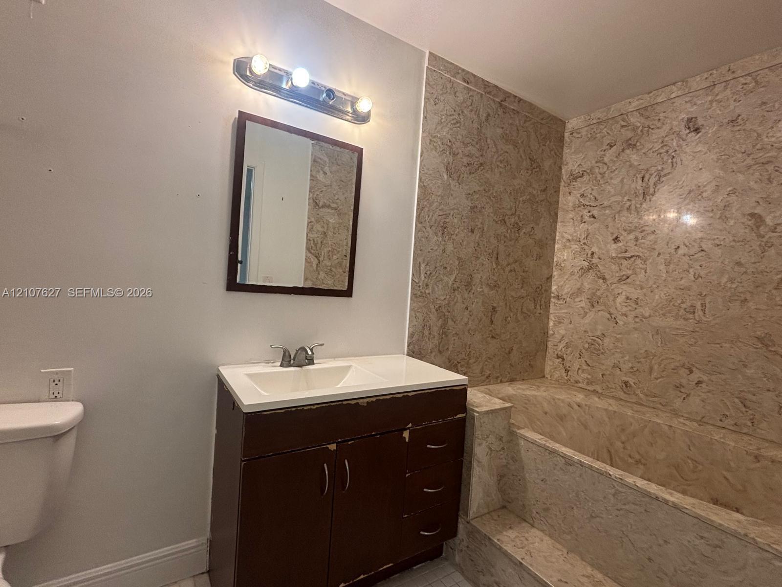 Bathroom featuring a vanity with an integrated sink, three-light wall-mounted fixture, framed mirror, and a tub surround with stone-patterned paneling