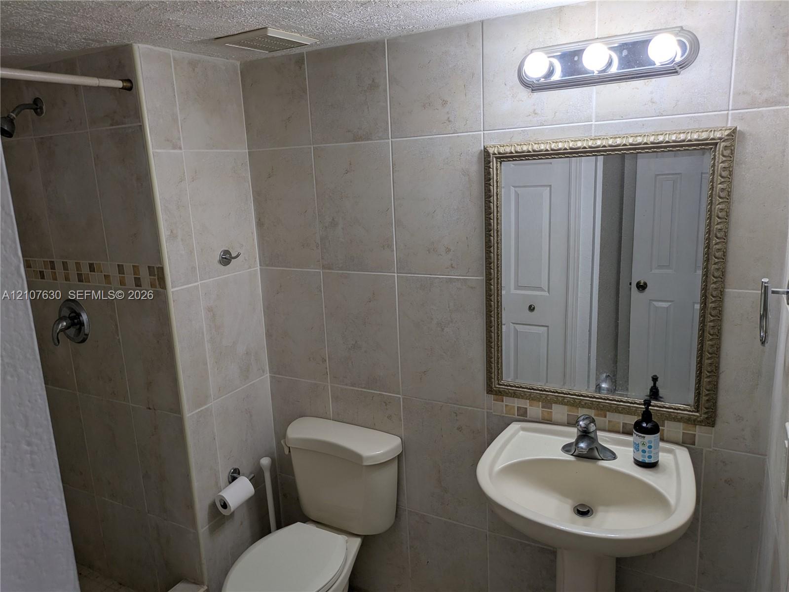 Bathroom featuring beige wall tiles with a glass mosaic accent band, a white pedestal sink with a chrome faucet, a framed wall mirror, and a walk-in shower