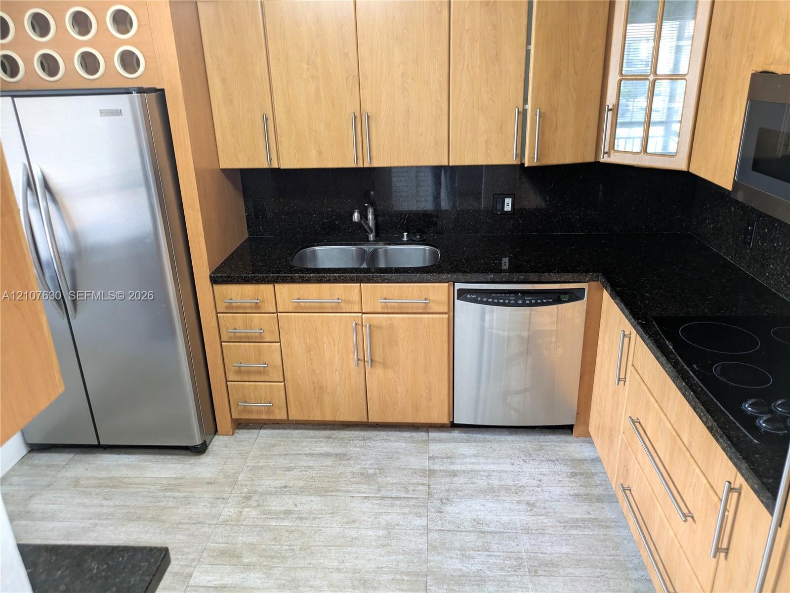 Kitchen featuring wood-finish cabinetry, dark stone countertops, stainless steel appliances, dual-basin stainless steel sink, and light-toned tile flooring