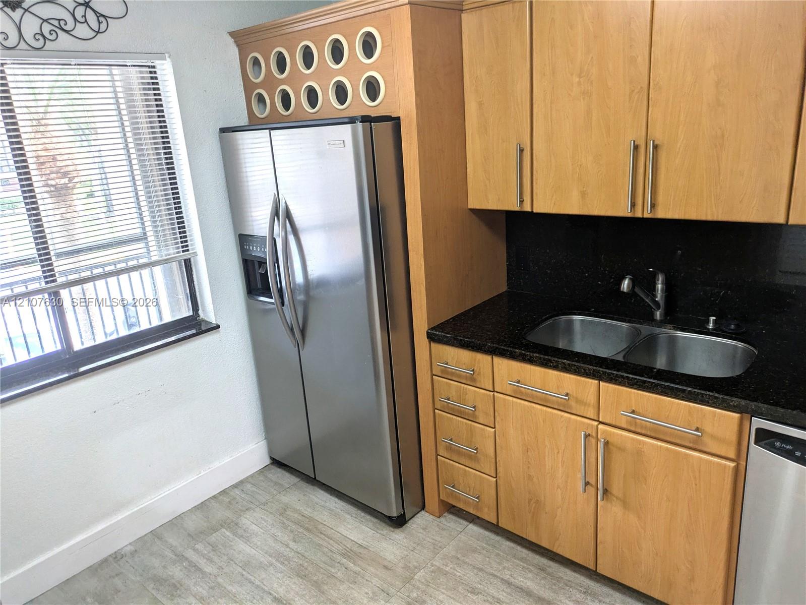 Kitchen featuring light wood-finish cabinetry, dark solid-surface countertops, dual-basin stainless steel sink with pull-down faucet, stainless steel side-by-side refrigerator, and tile flooring