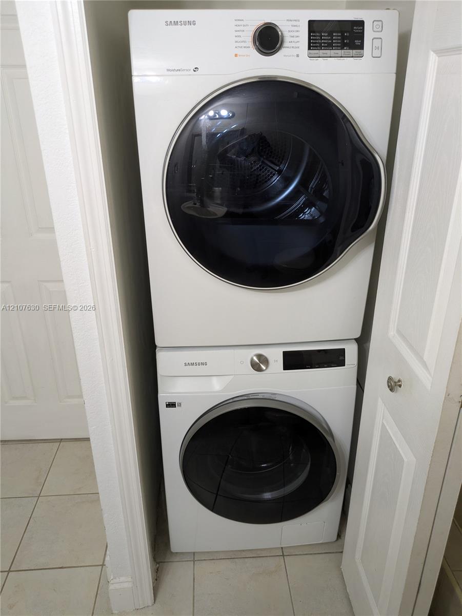 Stacked white front-load washer and dryer units with digital controls and glass-paneled doors, situated within a recessed laundry closet featuring tile flooring
