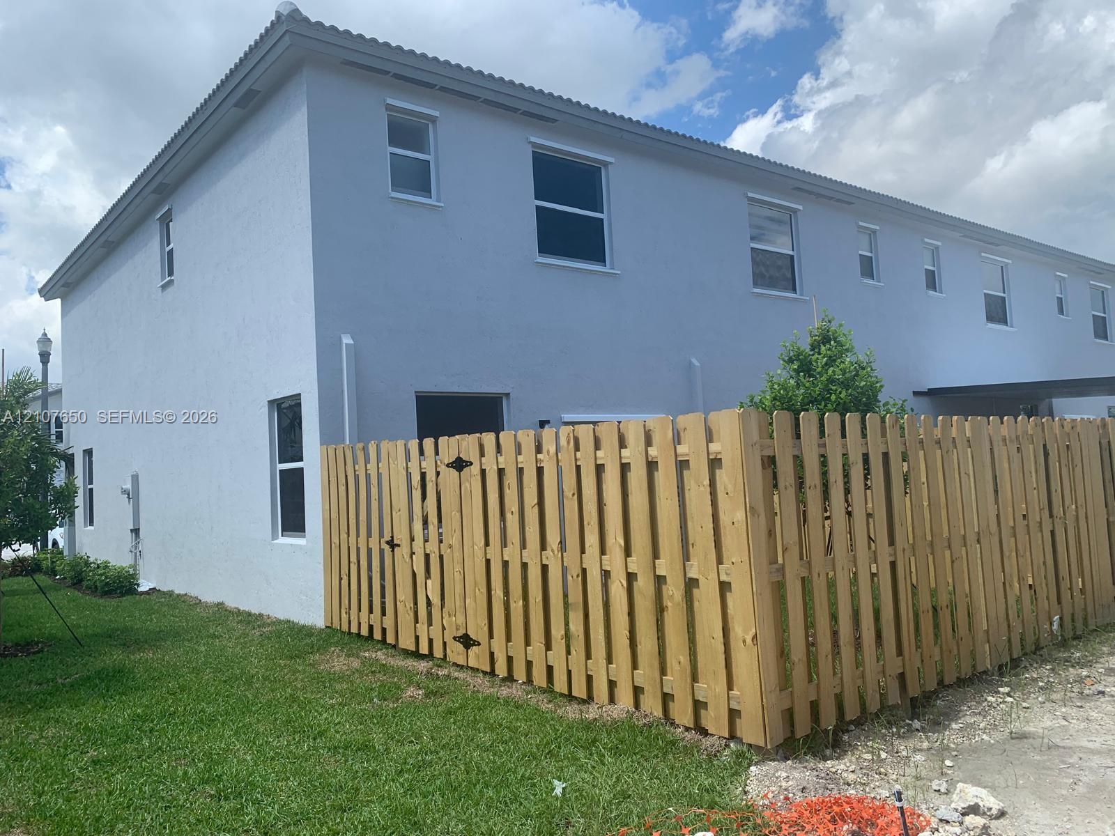 Two-story exterior with a light-toned stucco finish, multiple single-hung windows, and a wood-slat privacy fence enclosing a grass lawn