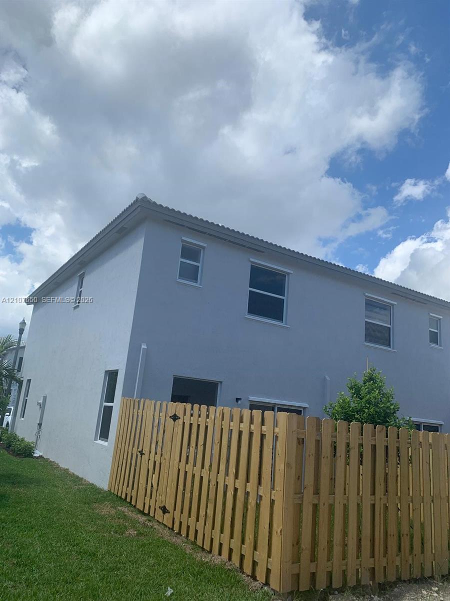 Two-story exterior featuring a light-toned stucco facade, neutral-hued tiled roof, multiple rectangular windows, and a wood-slat privacy fence